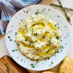 creamy whole30 mashed potatoes in white bowl with ghee on top next to cutting board, potatoes and rosemary