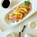 salmon tataki on plate garnished with lemon onion and sprouts next to chopsticks and ponzu