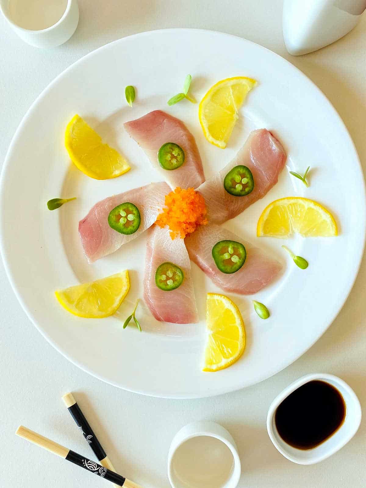 yellowtail sashimi slices on white plate topped with jalapeno slices and surrounded by lemon slices, soy sauce and chopsticks