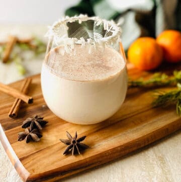 a glass of coquito with a coconut rim on a wood cutting board surrounded by spices, oranges and rosemary