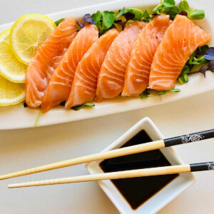 salmon sashimi on a bed of microgreens garnished with lemon next to soy sauce and chopsticks