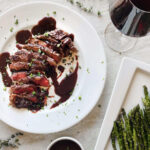 wagyu ribeye steak covered in wagyu bacon bordelaise sauce next to glass of red wine and asparagus