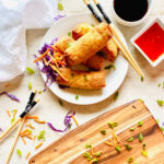 frozen egg rolls cooking in air fryer on a plate next to chopstick, soy sauce and thai chili dipping sauce