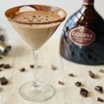 godiva chocolate martini in a martini glass with a bottle of Godiva chocolate liqueur, a cocktail shaker and chocolate in the background