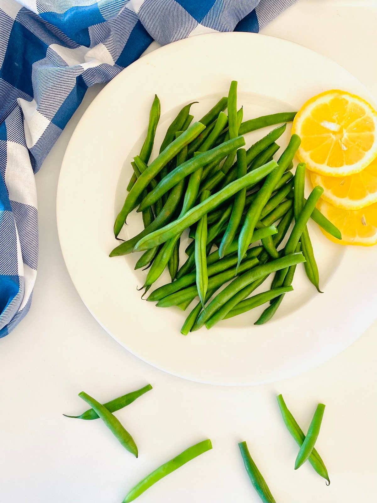 green beans on a plate next to lemon slices and a blue checkered cloth napkin
