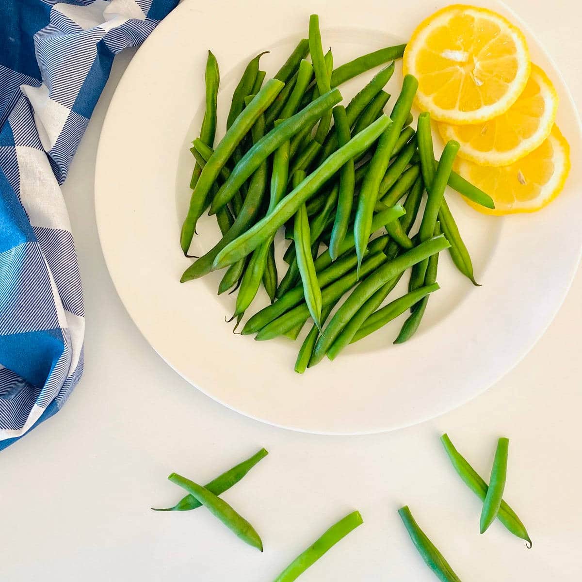 green beans on a plate next to lemon slices and a blue checkered cloth napkin