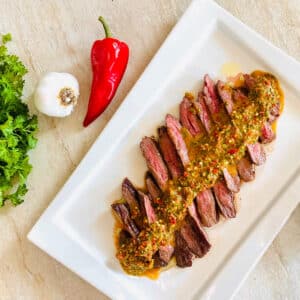 red chimichurri on top of sliced steak next to red pepper, garlic and cilantro