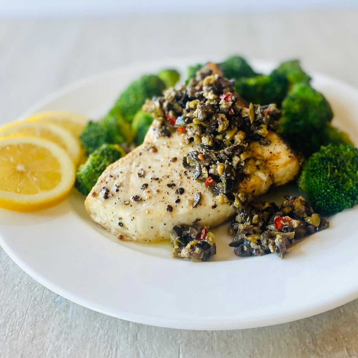 air fryer swordfish on a plate with broccoli, lemon slices and topped with olive tapenade