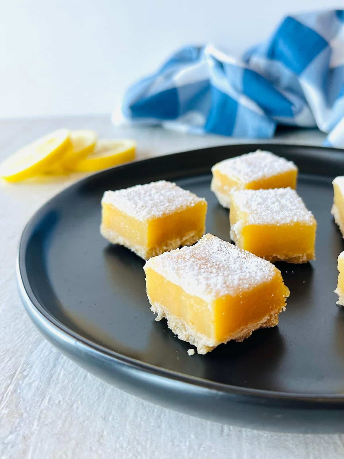 lemon cream cheese bars with graham cracker crust on black plate with sliced lemons and a blue napkin.