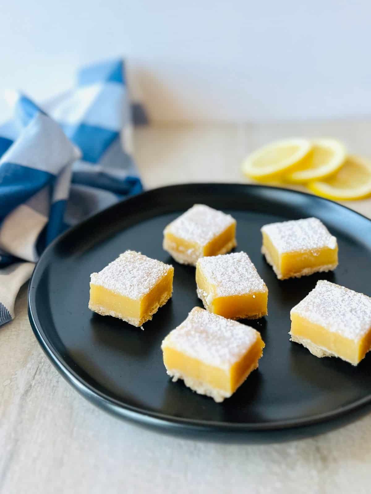 lemon squares with graham crust on black plate with sliced lemons and a blue napkin.