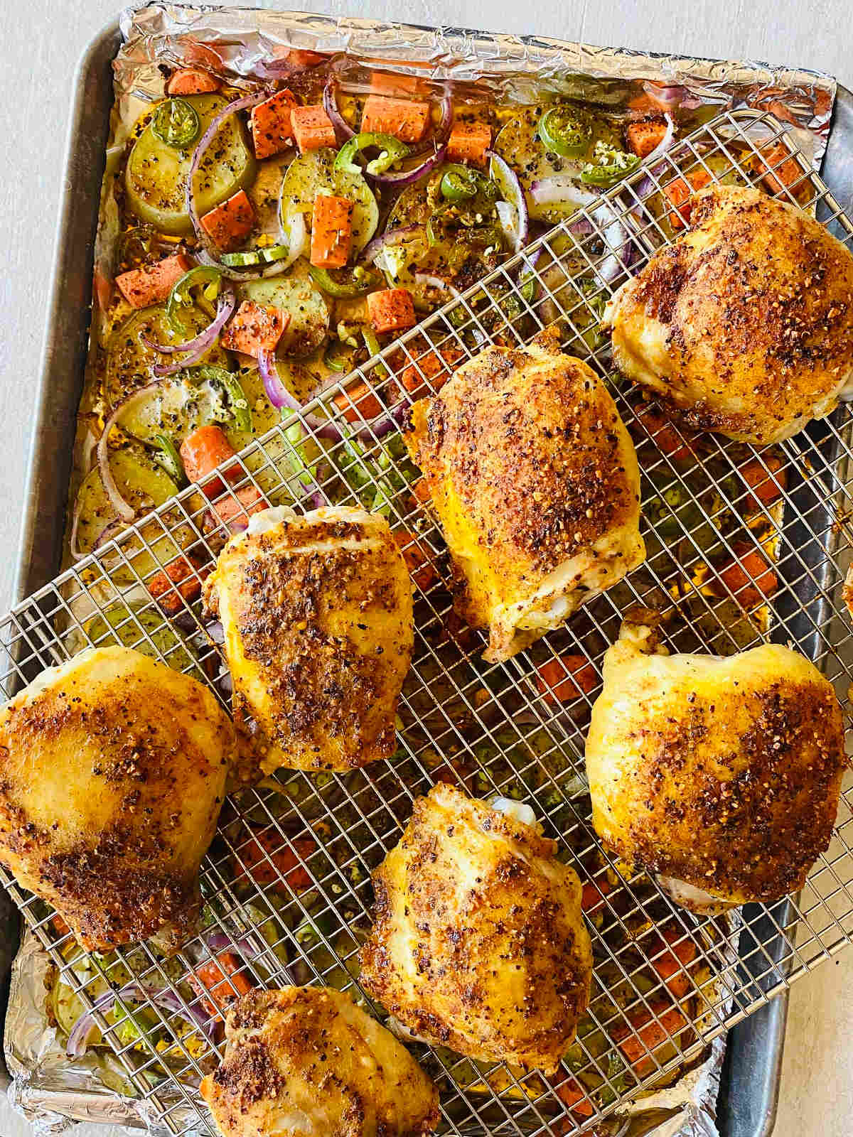sheet pan chicken thighs on a sheet pan over a baking rack with potatoes, carrots onions and jalapenos underneath.