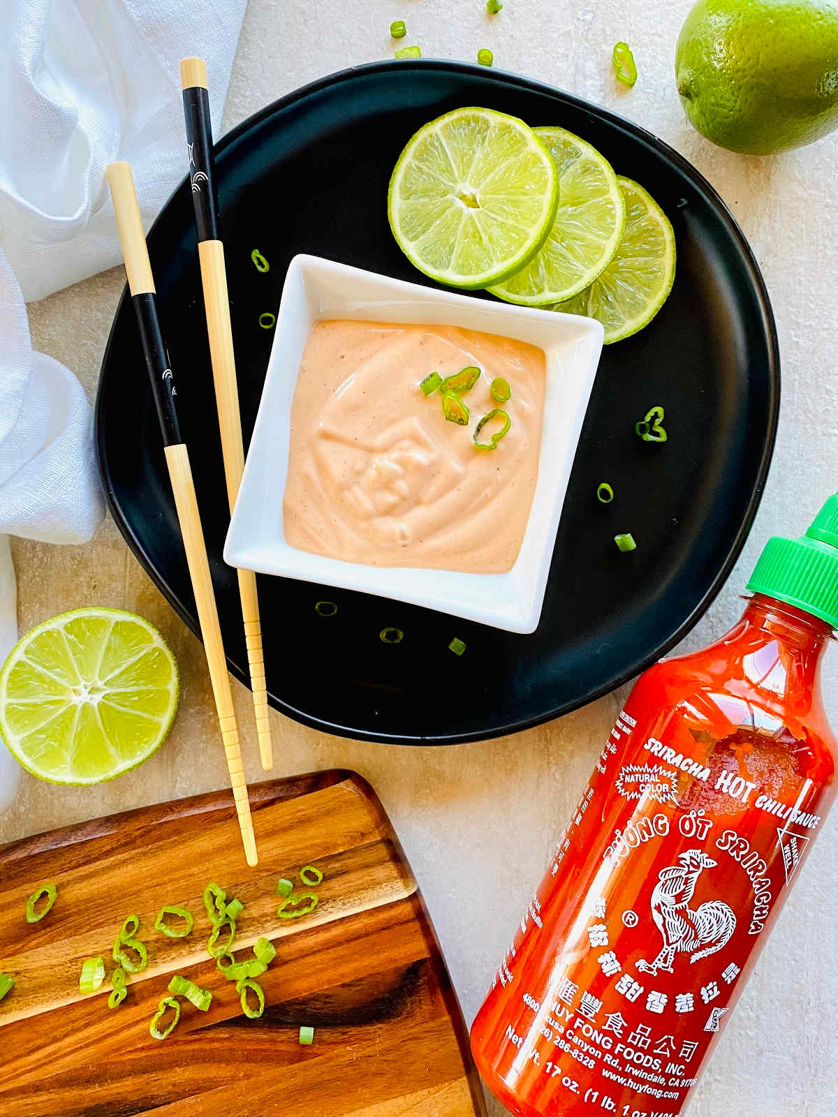 sriracha aioli in a dish next to a bottle of Huy Fong Sriracha Hot Chile Sauce with limes, chopsticks and scallions.