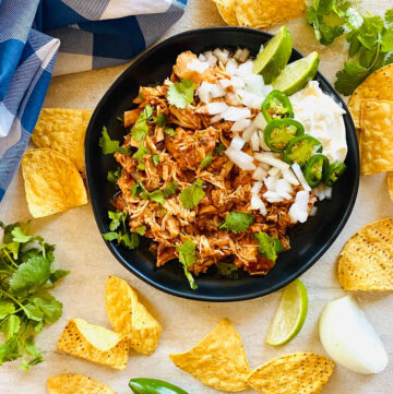 instant pot salsa chicken shredded, on a black plate, garnished with cilantro, diced onion, sliced jalapeno, sour cream and lime wedges. Tortilla chips are surrounding the plate.