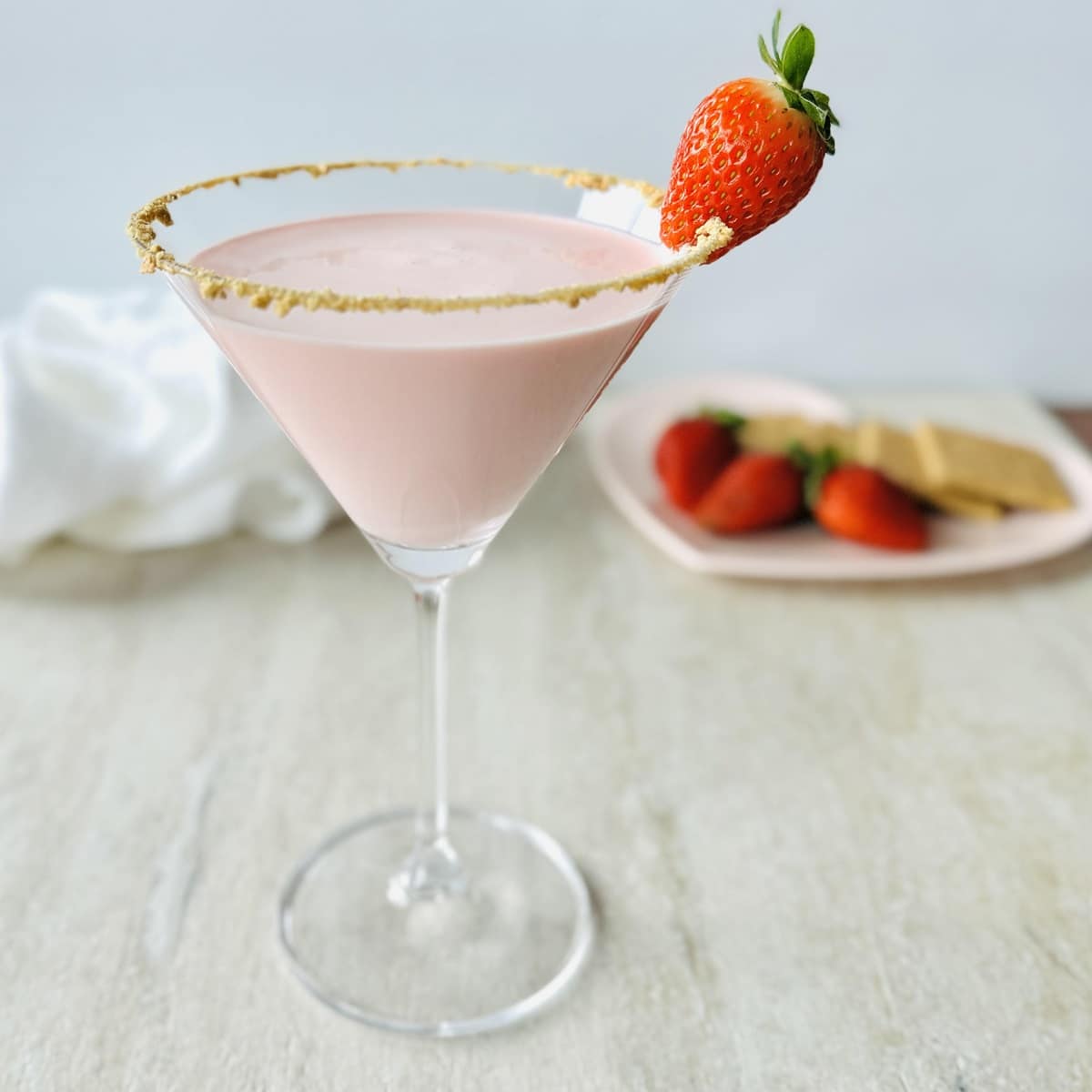 creamy strawberry cheesecake martini recipe in a martini glass with a graham cracker rim and fresh strawberry garnish.